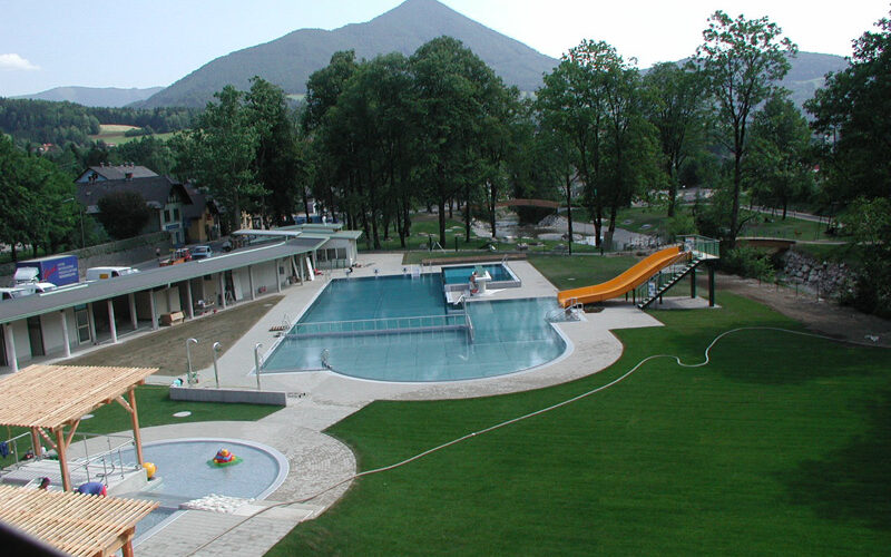 Freibadkomplex mit gelber Rutsche, grünem Rasen, umliegenden Bäumen und Bergen im Hintergrund unter einem klaren Himmel.