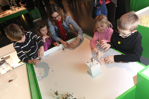 Ein Erwachsener und vier Kinder interagieren mit kleinen Objekten auf einem weißen Tisch in einem hell erleuchteten, modernen Ausstellungsraum.