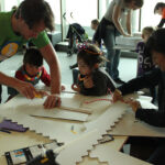 In einem hell erleuchteten Raum sitzen Erwachsene und Kinder an einem Tisch und arbeiten mit Papier, Lineal und Schere an Bastelprojekten.