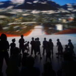 Eine schemenhafte Gruppe von Kindern und Erwachsenen steht und sitzt vor einer großen, verschwommenen Projektion einer Landschaftsszene.