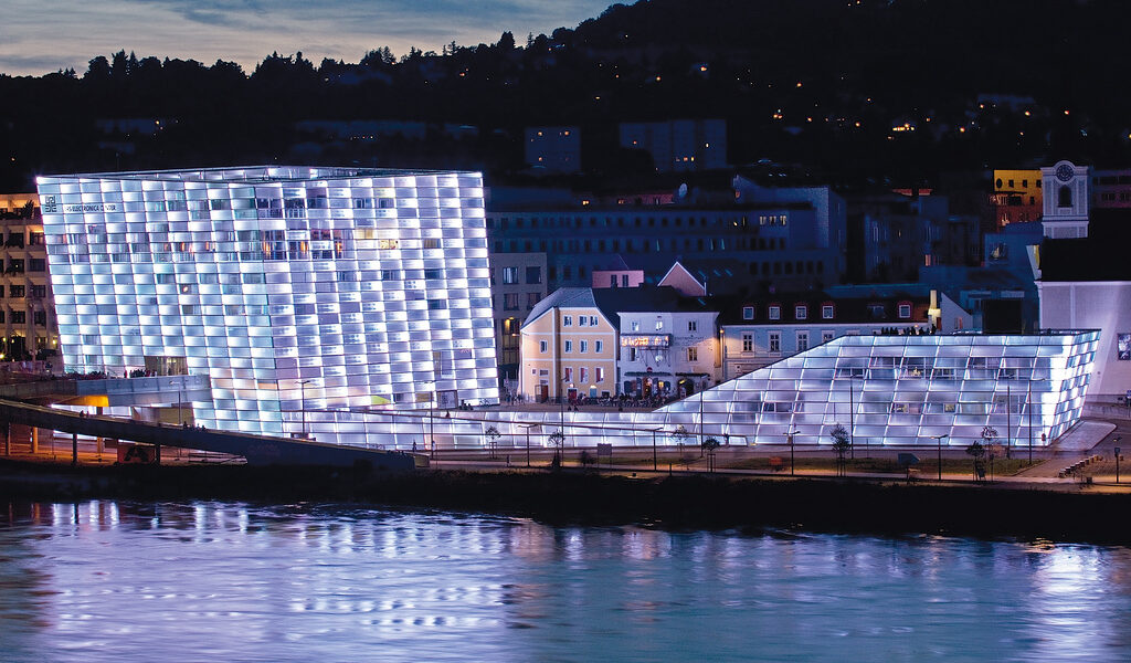 Ein modernes, mit weißem Licht beleuchtetes Gebäude mit Glasfront steht in der Abenddämmerung neben einem Fluss, im Hintergrund sind kleinere Gebäude und Bäume zu sehen.