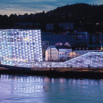 Ein modernes, mit weißem Licht beleuchtetes Gebäude mit Glasfront steht in der Abenddämmerung neben einem Fluss, im Hintergrund sind kleinere Gebäude und Bäume zu sehen.