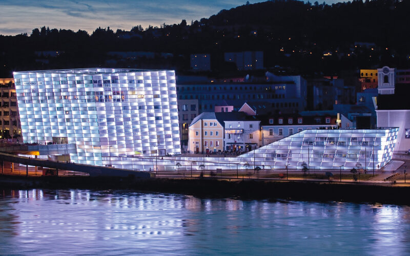 Ein modernes, mit weißem Licht beleuchtetes Gebäude mit Glasfront steht in der Abenddämmerung neben einem Fluss, im Hintergrund sind kleinere Gebäude und Bäume zu sehen.