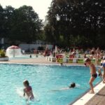 Kinder und Erwachsene erfreuen sich an einem sonnigen Tag an einem belebten Freibadbereich, in dem einige Kinder im Wasser spielen und andere sich darauf vorbereiten, ins Wasser zu springen.