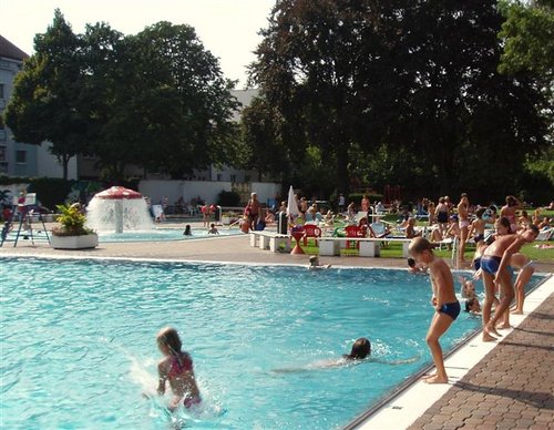 Kinder und Erwachsene erfreuen sich an einem sonnigen Tag an einem belebten Freibadbereich, in dem einige Kinder im Wasser spielen und andere sich darauf vorbereiten, ins Wasser zu springen.