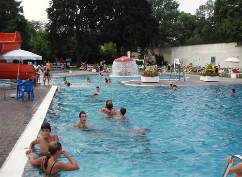 Der Außenpool mit Wasserfontäne, Rutsche und Liegestühlen, umgeben von Bäumen und Grün, lädt zum Schwimmen und Entspannen ein.