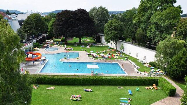 Ein großer Freibadkomplex mit Rutsche, umgeben von Gras, Bäumen und Menschen, die sich sonnen oder auf Liegestühlen entspannen.