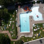 Luftaufnahme eines öffentlichen Schwimmbads mit schwimmenden und sich sonnenden Menschen auf Liegestühlen auf der Wiese.
