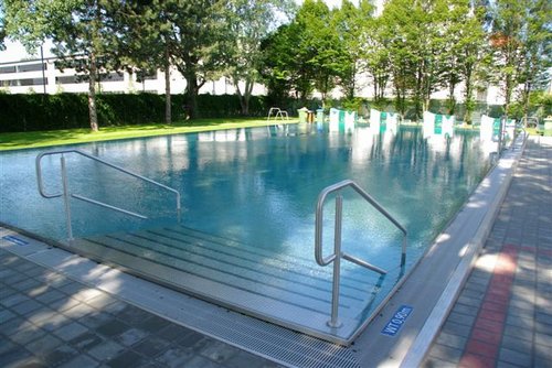 Außenschwimmbad mit Stufeneinstieg und Handläufen, umgeben von Bäumen und einer gepflasterten Fläche; im Hintergrund sind Schwimmbadschilder und Stühle zu sehen.
