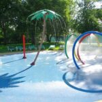 Ein Planschbecken mit Wasserspielen, darunter eine Palme, bunte Ringe und Bodenfontänen, umgeben von Liegestühlen und Bäumen.