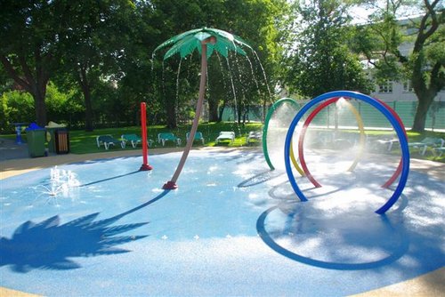 Ein Planschbecken mit Wasserspielen, darunter eine Palme, bunte Ringe und Bodenfontänen, umgeben von Liegestühlen und Bäumen.