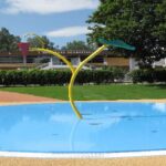 Ein kleines Planschbecken mit seichtem, blauem Wasser, zwei gelbe Sprühvorrichtungen in Form von Blättern und ein Schulgebäude im Hintergrund an einem sonnigen Tag.