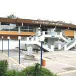 Ein modernes, rechteckiges Gebäude mit der Bezeichnung "Hallenbad Donaustadt" mit Außentreppe, großen Fenstern und einem gepflasterten Eingang mit Laternenpfählen und einer Skulptur.