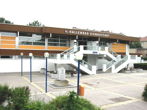 Ein modernes, rechteckiges Gebäude mit der Bezeichnung "Hallenbad Donaustadt" mit Außentreppe, großen Fenstern und einem gepflasterten Eingang mit Laternenpfählen und einer Skulptur.