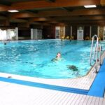 Hallenbad mit klarem, blauem Wasser; ein Schwimmer ist im Becken, und im Hintergrund sind einige Personen auf dem Pooldeck zu sehen.