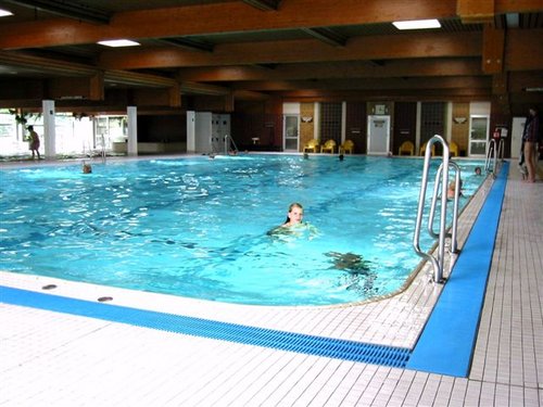 Hallenbad mit klarem, blauem Wasser; ein Schwimmer ist im Becken, und im Hintergrund sind einige Personen auf dem Pooldeck zu sehen.