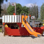 Eine Spielplatzstruktur in Form eines rot-weißen Schiffes mit einer orangefarbenen Rutsche, die auf einer Kiesfläche im Freien steht.
