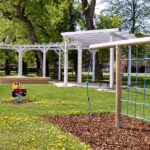 Ein Spielplatz mit Klettergeräten, einem Springreiter und einer weißen Pergola in einem Park mit Bäumen und Gras.
