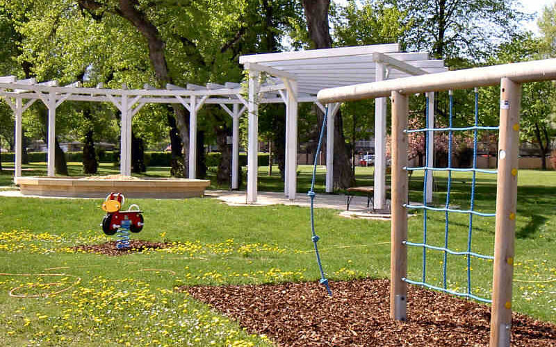Ein Spielplatz mit Klettergeräten, einem Springreiter und einer weißen Pergola in einem Park mit Bäumen und Gras.