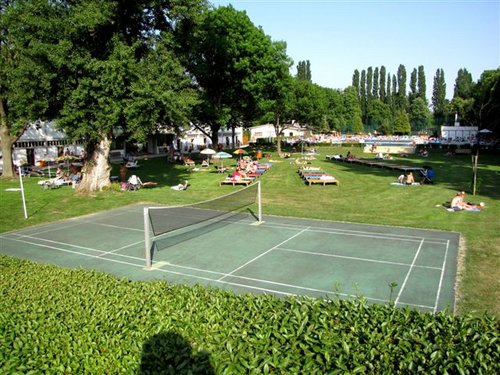 Badmintonplatz im Freien auf einer Wiese mit Sonnenbadenden und Entspannern auf Liegestühlen im Hintergrund, umgeben von Bäumen.