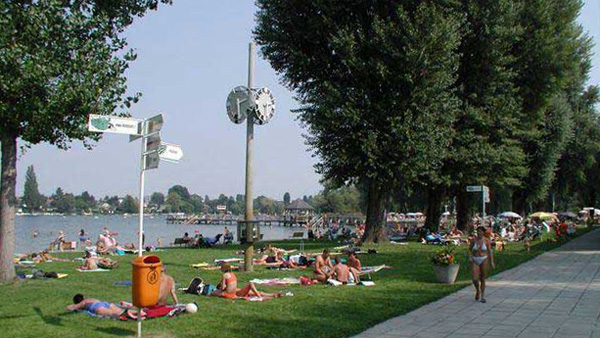 Menschen, die sich auf einer Wiese am See sonnen und entspannen, mit Bäumen, einem Abfalleimer, Schildern und einer großen Uhr; im Hintergrund Wasser und entfernte Gebäude.