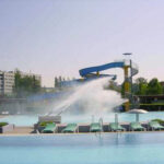 Ein Pool mit Liegestühlen, einer großen Wasserrutsche und einem Wasserspiel in der Mitte. Im Hintergrund sind Wohnhäuser und Bäume zu sehen.