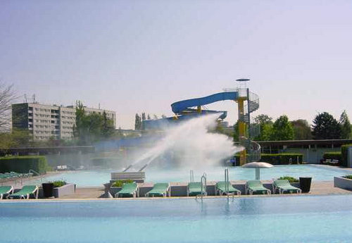 Ein Pool mit Liegestühlen, einer großen Wasserrutsche und einem Wasserspiel in der Mitte. Im Hintergrund sind Wohnhäuser und Bäume zu sehen.