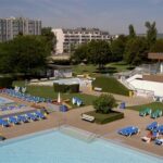 Freibadkomplex mit mehreren Becken, zahlreichen blauen Liegestühlen, ein paar Sonnenbadenden und Wohngebäuden im Hintergrund.