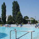 Öffentliches Freibad mit Menschen, die schwimmen und sich entspannen, umgeben von Bäumen und Gebäuden an einem sonnigen Tag.