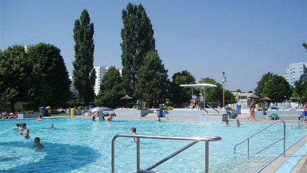 Öffentliches Freibad mit Menschen, die schwimmen und sich entspannen, umgeben von Bäumen und Gebäuden an einem sonnigen Tag.