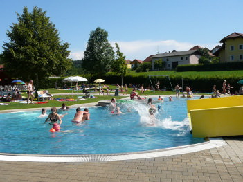 Menschen schwimmen und spielen an einem sonnigen Tag in einem Freibad, andere entspannen sich auf der nahe gelegenen Wiese; im Hintergrund sind Bäume und Häuser zu sehen.
