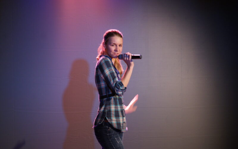 Eine Person in einem karierten Hemd hält ein Mikrofon in der Hand und performt auf der Bühne unter rosa und blauer Beleuchtung.