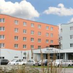 Ein modernes Hotelgebäude mit pfirsichfarbener und hellblauer geschwungener Fassade, mehreren Fenstern, geparkten Autos davor und einem teilweise bewölkten Himmel darüber.