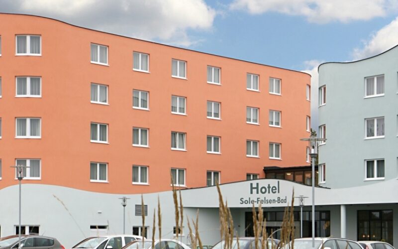 Ein modernes Hotelgebäude mit pfirsichfarbener und hellblauer geschwungener Fassade, mehreren Fenstern, geparkten Autos davor und einem teilweise bewölkten Himmel darüber.