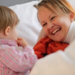 Eine Frau lächelt, während sie auf einem Bett neben einem kleinen Kind liegt, das ein rosa kariertes Hemd trägt.