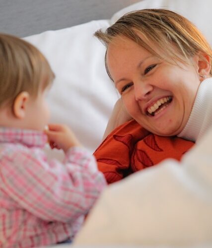 Eine Frau lächelt, während sie auf einem Bett neben einem kleinen Kind liegt, das ein rosa kariertes Hemd trägt.