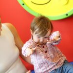 Ein Erwachsener hält ein Kleinkind in einem karierten Hemd, das seine Arme nach vorne streckt. Der Hintergrund ist leuchtend rot, wobei ein Teil eines gelben, runden Objekts sichtbar ist.