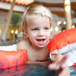 Ein kleines Kind mit blondem Haar, das orangefarbene aufblasbare Armbänder trägt, lächelt, während es in einem Schwimmbad spielt.