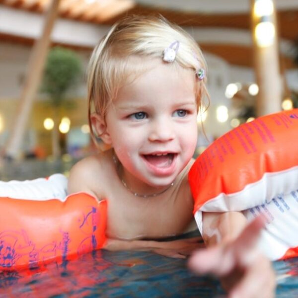 Ein kleines Kind mit blondem Haar, das orangefarbene aufblasbare Armbänder trägt, lächelt, während es in einem Schwimmbad spielt.