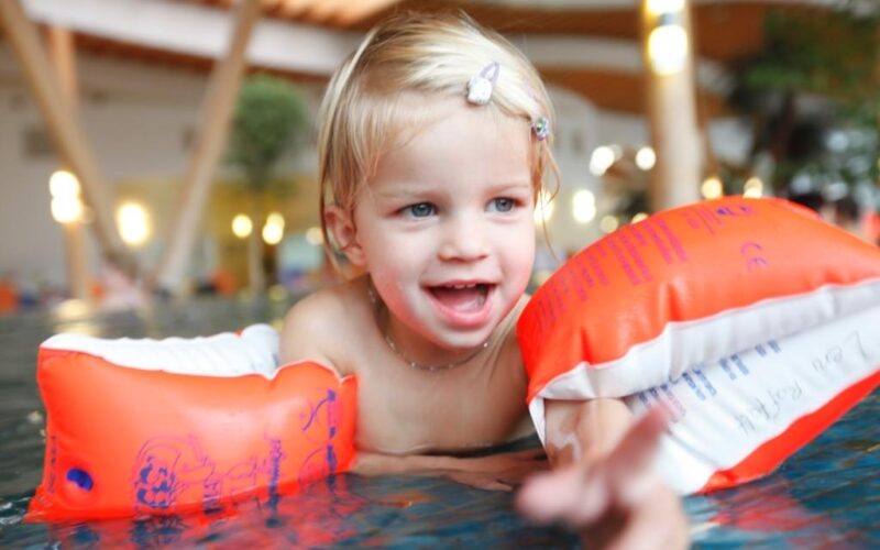 Ein kleines Kind mit blondem Haar, das orangefarbene aufblasbare Armbänder trägt, lächelt, während es in einem Schwimmbad spielt.