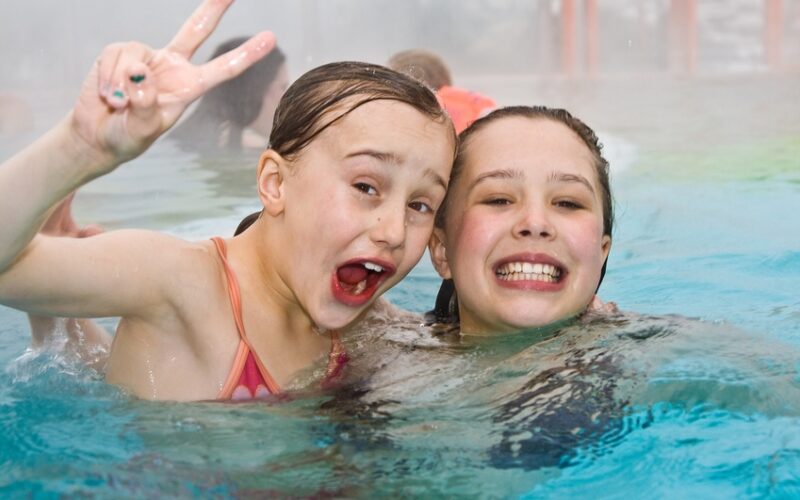 Zwei Mädchen lächeln und posieren in einem Swimmingpool, wobei eines ein Friedenszeichen macht und beide in die Kamera schauen.