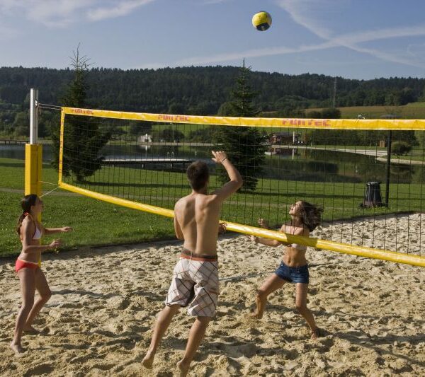 Drei Personen spielen Beachvolleyball auf einem Sandplatz in einem Park mit Gras, Wasser und Bäumen im Hintergrund unter blauem Himmel.