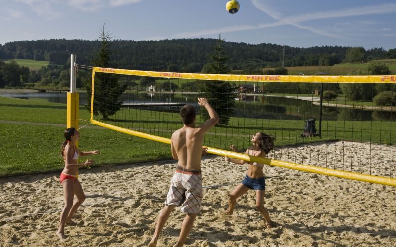 Drei Personen spielen Beachvolleyball auf einem Sandplatz in einem Park mit Gras, Wasser und Bäumen im Hintergrund unter blauem Himmel.