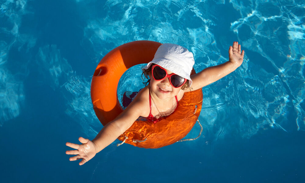 Kind mit roter Sonnenbrille und weißer Mütze in einem orangefarbenen Schwimmring, lächelnd und mit ausgebreiteten Armen, während es in einem Schwimmbecken schwimmt.