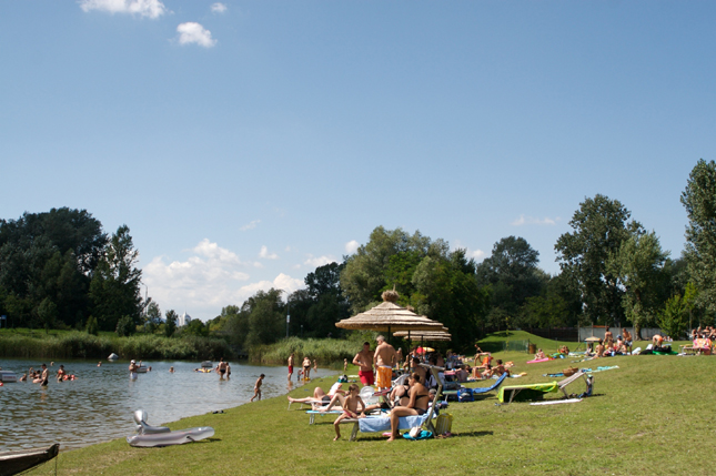 An einem sonnigen Tag entspannen sich die Menschen auf Liegestühlen und schwimmen in einem kleinen, von Bäumen umgebenen See, der von einem strohgedeckten Sonnenschirm beschattet wird.