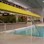 Hallenbad mit Metallgeländern, gefliestem Boden und einem gelben Balkon darüber. An der Seite befinden sich Pflanzen und ein Verkehrskegel in der Nähe des Beckenrandes.