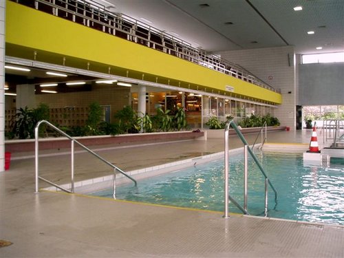 Hallenbad mit Metallgeländern, gefliestem Boden und einem gelben Balkon darüber. An der Seite befinden sich Pflanzen und ein Verkehrskegel in der Nähe des Beckenrandes.