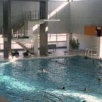 Hallenbad mit mehreren schwimmenden Personen und einem großen Sprungturm im Hintergrund. Das Sonnenlicht strömt durch die oberen Fenster ein und erhellt den Raum.