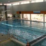 Hallenbad mit Bahntrennern, Sprungbrettern und großen Fenstern, die natürliches Licht hereinlassen; ein Verkehrskegel befindet sich in der Nähe des flachen Endes.