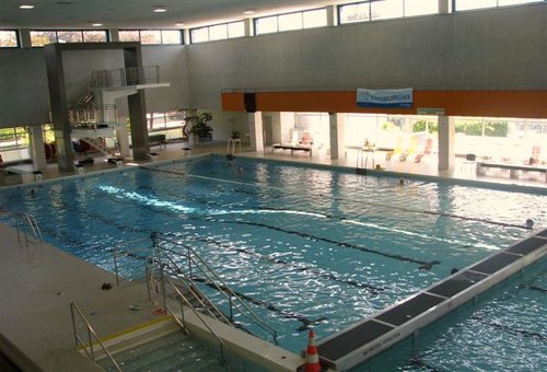 Hallenbad mit Bahntrennern, Sprungbrettern und großen Fenstern, die natürliches Licht hereinlassen; ein Verkehrskegel befindet sich in der Nähe des flachen Endes.